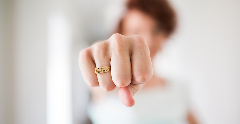 A woman in a white short holds her fist out determinedly toward the camera. She wears a metal ring stamped with the words "I am bad ass"