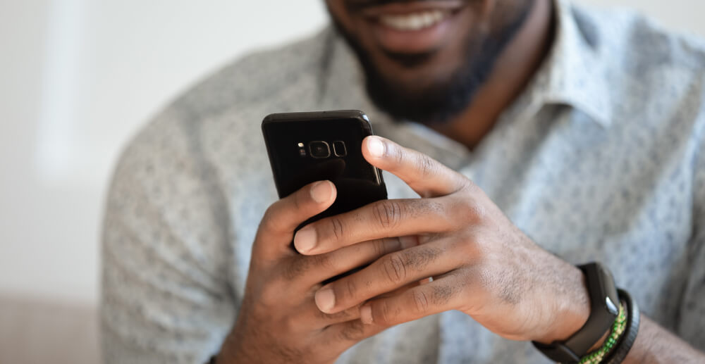 A Black man smiles as he uses a smartphone.