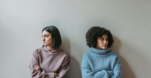 Two young women sit side by side, both with their arms crossed. They are facing away from one another with disgruntled expressions.