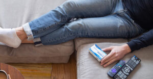 A person wearing blue jeans sits on a sofa with tehir legs stretched out in front of them, a box of Suboxone by their side.