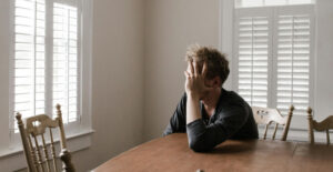 A man with blonde hair sits at a dining table. He leans one elbow on the talbe and holds his face in his hand, in a posture of distress.