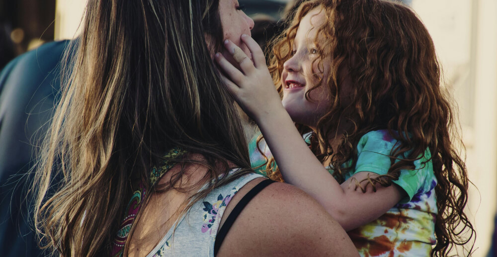 A woman in a tank top holds a little girl with red curls. The girl is smooshing the woman's face between her little hands.