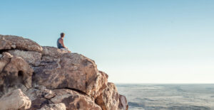 A person sits alone on a rocky outcropping, staring at the sea.