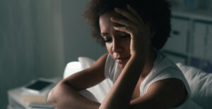 An unhappy Black woman sits on a bed, with one hand held up to her head in a sad pose.