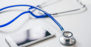 A blue stethoscope and a smartphone lie on a white desk.