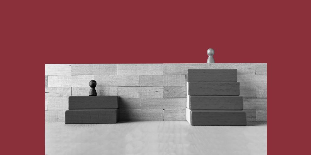 Wooden game pieces representing inequality. A dark piece stands on two stair-step blocks, facing the barrier of a high wooden wall. The light piece stands on top of the wall, with four stair-step blocks leading the way up.