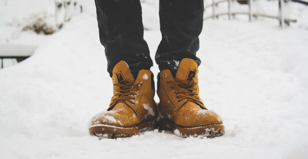 A person's feet in brown hiking boots, standing in the snow.