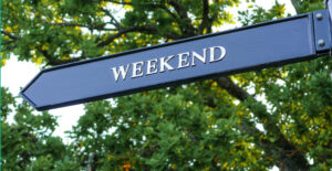 A metal street sign that reads "Weekend," n front of leafy green tree branches.