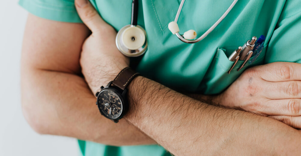 A doctor in mint green scrubs stands with his arms crossed. The camera does not show his face.