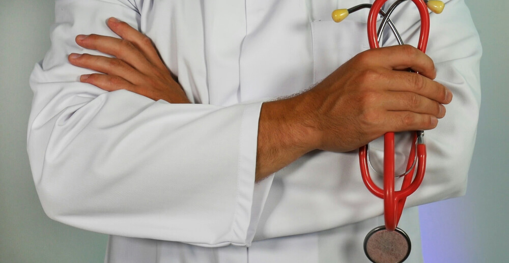 A male doctor in a white lab coat stands with his arms crossed, holding a stethoscope.