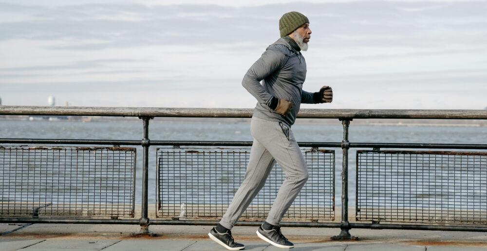 A middle-aged Black man with a gray beard wears a track suit and a knit cap as he runs along a city seaside.