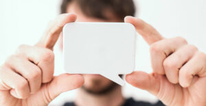 A man with short brown hair holds a white speech bubble cutout in front of his face, up to the camera.