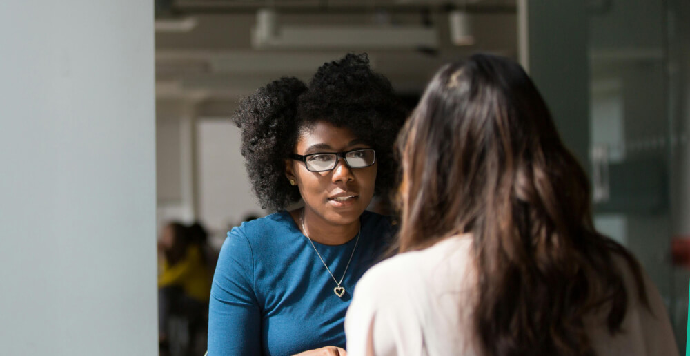 Two young women sit across the table from one another in coversation. One is Black, the other White.