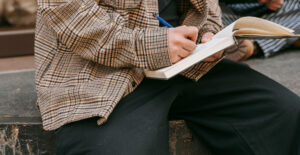 A person in a plaid shirt balances a journal on their knee and writes in it.