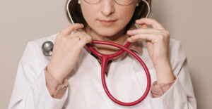 A female doctor in a white lab coat lifts the earpieces of a stethoscope into place