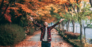 A man in a red plaid shirt stands with autumn trees behind him, throwing leaves in the air with a big smile on his face