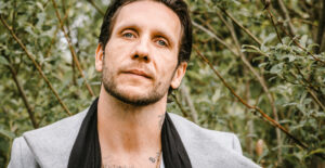 Brandon Novak wears a fashionable sportscoat while sitting outdoors.