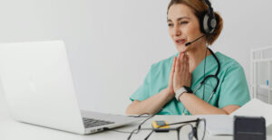 A female doctor in scrubs uses a laptop to have a video appointment with her patient.