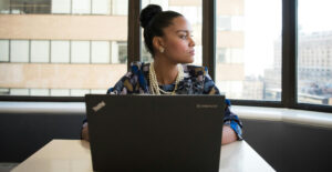 A woman in professional clothes sits at a desk with a laptop on it and stares pensively out the window.