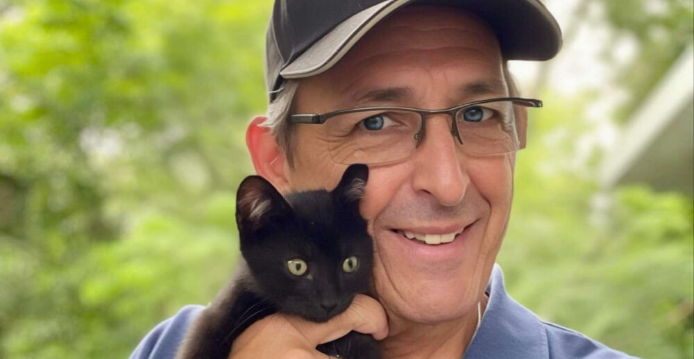 Tim is a man in glasses and a baseball cap, holding a small and adorable black cat.