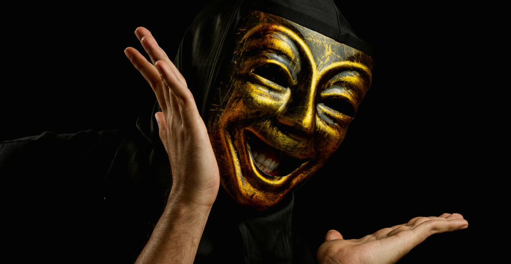 In a dark space, a person wears a gleaming golden comedy mask and gestures widely with their arms.