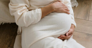 Torso of a pregnant woman in a white sweater dress, with her hands gently holding her rounded belly