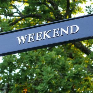 A metal street sign that reads "Weekend," n front of leafy green tree branches.