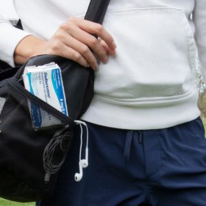 A man in a sweatshirt stands outdoors, carrying a gym bag. Visible through the mesh pocket of the bag is a box of Suboxone.