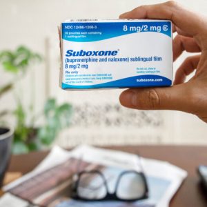 A hand holds a slightly battered box of Suboxone up to the camera. Beyond the box is a table holding a newspaper, a pair of glasses, and a mug.