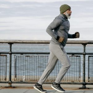 A middle-aged Black man with a gray beard wears a track suit and a knit cap as he runs along a city seaside.