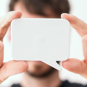 A man with short brown hair holds a white speech bubble cutout in front of his face, up to the camera.