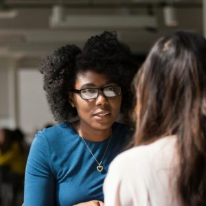 Two young women sit across the table from one another in coversation. One is Black, the other White.