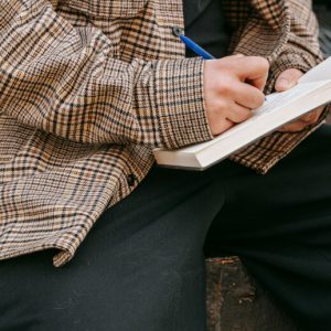 A person in a plaid shirt balances a journal on their knee and writes in it.