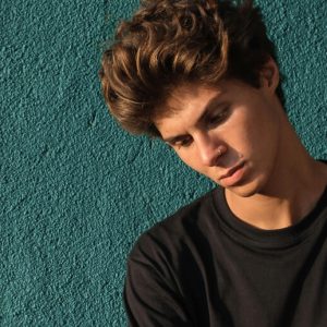 A young man looks down pensively. He sits in front of a turquoise wall.