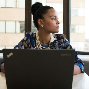 A woman in professional clothes sits at a desk with a laptop on it and stares pensively out the window.