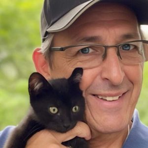 Tim is a man in glasses and a baseball cap, holding a small and adorable black cat.