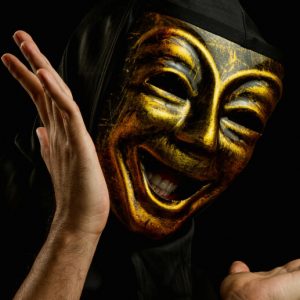 In a dark space, a person wears a gleaming golden comedy mask and gestures widely with their arms.