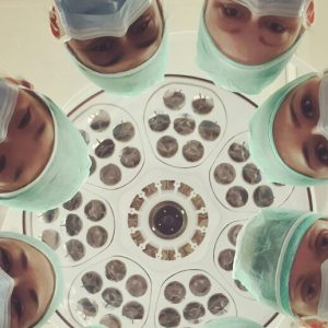 Several doctors, masked for surgery, stand in a circle, staring down at the camera.