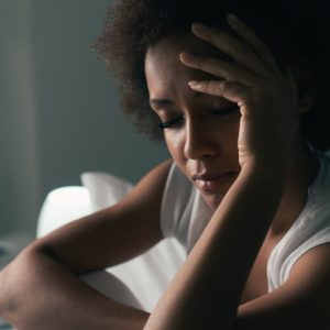 An unhappy Black woman sits on a bed, with one hand held up to her head in a sad pose.