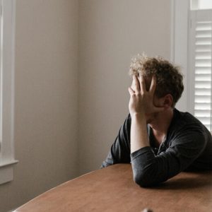 A man with blonde hair sits at a dining table. He leans one elbow on the talbe and holds his face in his hand, in a posture of distress.