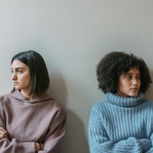 Two young women sit side by side, both with their arms crossed. They are facing away from one another with disgruntled expressions.